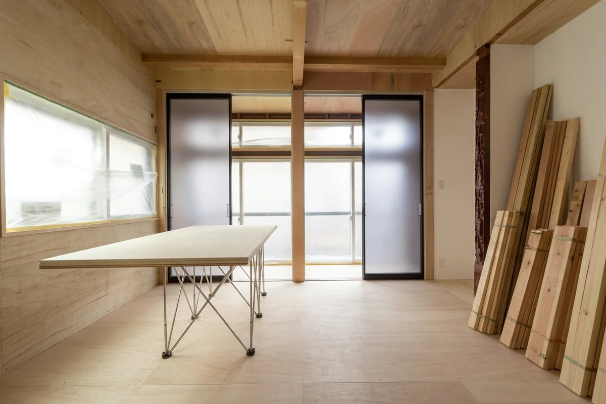 工事中の部屋。木の天井と合板の床、窓の養生、壁ぎわに束ねて立てかけた無垢材と作業台(所沢・ねこねすとの現場)